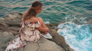 Gürültülü video. Genç bir kadının silueti. Kadın öfkeli mavi denizin taştan sahilinde oturuyor, dalgalar kayalara çarpıyor. Arkadan bak. Costa Brava plajındaki mutlu kız turist Lloret de Mar