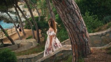 Neşeli mutlu genç kadın turist Lloret de Mar İspanya parkında yürüyor. Arkadan bakan romantik seksi kız. Costa Brava yaz doğası yeşil ağaç bahar günbatımı. Uzun, zarif, klasik, güneşli elbise.