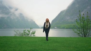 Mutlu kadın turist yağmurda Hallstatter Gölü yakınlarında yeşil çimlerde yürüyor. Alplerdeki kız, bulutlardaki dolomite dağları, sis, dramatik gökyüzü. Turizm köyü Hallstatt. Soğuk yağmurlu bahar. Bir kişi.