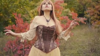 Fantezi kadın ortaçağ cadısı el kaldırarak dua ediyor. Eski Bromn tarzı elbise. Kız prenses sonbahar portakal yeşilliği ormanının doğasında duruyor. Yaratıcı kraliyet giysisi, klasik elbise. Uzun kızıl saçlı.