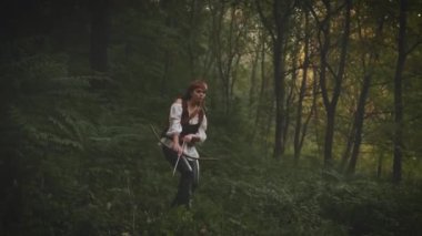 Gürültülü video. Kadın ormanda yürüyor. Kızın elinde ahşap yay var. Lady Hunter 'ın fantezi görüntüsü. Kaliteli savaşçı okçuluk. Deri pantolon, beyaz gömlek, oklu çanta.