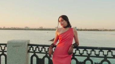 Uzun kırmızı elbiseli zarif bayan Dnipro nehrinin setinde duruyor. Akşam makyajı seksi surat. Şık kadın tül eldivenleri. Boynundaki ipek eşarp rüzgarda uçuşuyor. Kadınlar gün batımını sever