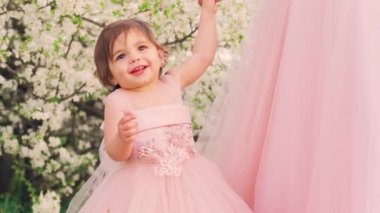 Portre. Küçük mutlu, neşeli, neşeli kız kız kız. Annesinin elini tutuyor. Pembe tüylü, kaliteli bir elbise. Çocuk prenses gülen surat. Arka planda bahar doğası çiçek açan beyaz ağaç yeşili çimenler