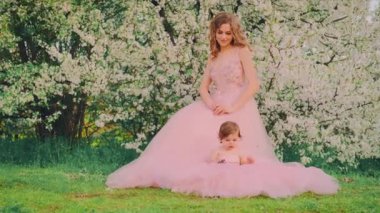 Fantezi portresi iki şirin mutlu kız kardeş prenses dinleniyor, küçük kız yeşil çimenlerde oturuyor ve oynuyor. Uzun pembe tül klasik ortaçağ kraliyet elbisesi. Bahar doğası beyaz çiçekler açan kiraz ağaçları