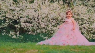Küçük mutlu, güzel, neşeli, şen şakrak pembe elbiseli kız portresi. Gülen yüzlü çocuk prenses balo elbisesi tarzı. Arka planda bahar doğası çiçek açan beyaz ağaç yeşili çimenler