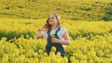 Yakın plan genç mutlu kız portresi. Kızıl saçlı gencin eli uzun saçları geri atıyor. Beyaz ceketli tulum. Çocuk gülüşü neşeyle güler. Arkaplan sarısı çiçek açan kolza tohumu baharı doğası