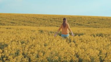Güzel, genç ve mutlu aktif kadın sarı çiçek açan kolza tohumu tarlasında koşuyor. Yazın mavi gökyüzü, alerji özgürlüğü konsepti. Saçlar rüzgarda yavaş çekimde uçar. Bahar çiçeği çayırı. arka görünüm