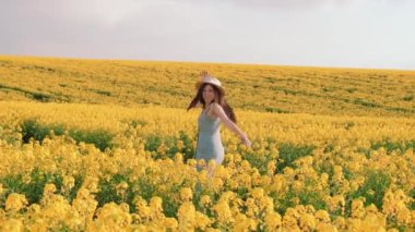 Neşeli kız. Dans eden mutlu, aktif genç bir kadın el ele tutuşuyor. Çiçekli bahar tarlası. Sarı kolza çiçeği. Kıllı esmer dalgalar rüzgarda uçuşuyor. Hasır şapka sundress. Mavi gökyüzü doğası