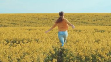 Genç ve güzel bir kadın kollarını açmış sahada koşuyor. Mutlulukla dans ederken sırt üstü eğlencenin görüntüsü. Sarı çiçek açan kolza tohumu. Sarışın, uzun saçlı. Turuncu ceketli, kot pantolonlu. Arkaplan mavi gökyüzü