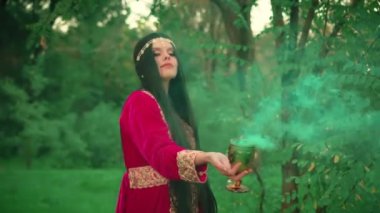 Gürültülü video. Fantezi kadın cadı aşk iksiri dumanıyla sihirli bardağı tutuyor. Kız kraliçe ormanda sihirbazlık yapıyor. Geleneksel kırmızı etnik türkçe elbise. Yeşil ağaçlar doğa