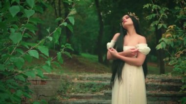 Gürültülü video. Eski tarz ortaçağ fantazisi kadın. Beyaz, uzun, eski bir elbise. Orman perisi kız. Bayan manken merdivenlerde poz veriyor. Yunan seksi tanrıçası çıplak omuzlar yeşil ağaç yaz doğası