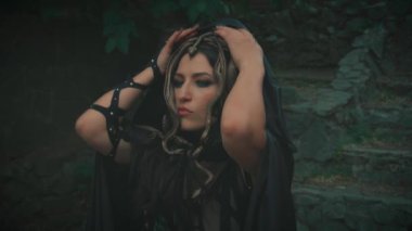 Gürültülü video. Cadılar Bayramı fantezi kadın Medusa gorgon Yunan tanrıçası Kara gotik kostüm. Kız pelerinini çıkarıyor. Başında altın yılanlar, korkunç yüz makyajı. Kara Kraliçe kötü cadı.