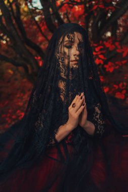 Sanat fotoğrafı gerçek gotik kadın siyah dantelli şal başörtüsü uzun saç örtüsü gizleme yüzü, eski tarzda giyinme. Vampir kadın seksi gözleri kapalı, elleri dua ederken katlanmış, sonbahar ormanı kırmızı ağacı