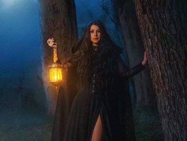 fotoğraf gerçek kişi, gotik kadın kraliçe karanlık, yoğun sis ormanlarında yürüyen eller fener lambasını mistik bir şekilde aydınlatıyor. Uzun siyah pelerinli kapüşon. Fantezi seksi kız Gece doğa ağaçları ay ışığı
