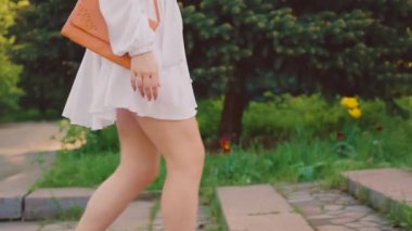 Yeşil çimen ağacında yürüyen mutlu kadın yaz mevsiminin tadını çıkarıyor. Beyaz ipek kısa elbise çantalı seksi manken kız çiçek açan bahçeye gider. Çiçek tutan el.