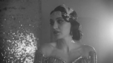 1920 'lerin siyah beyaz filmleri. Üzgün yüzlü genç retro kadın. Parlak, parlak, gümüş bir oda, parlak bir duman. Yakın plan portre kızı kafasında tüy aksesuarlarıyla parmak sallayan saç bandı.