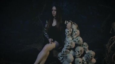 Gürültülü film. Cadılar Bayramı kadın vampir kostümü. Hayalet genç seksi gotik kadın. Yetişkin vampir kız kafatasındaki kurbanların tahtında oturuyor. Gece gotik karanlık orman. Siyah yaratıcı elbise. Seksi kötü kraliçe 4k