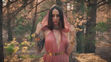 İmge Ormanı Büyücüsü Elf sihirbazının portresi. Seksi kız kameraya bakar ipek kapüşonlu pelerinini çıkarır seksi fantezi tarzı pembe elf elbisesi giyer. Sonbahar orman ağaçları.