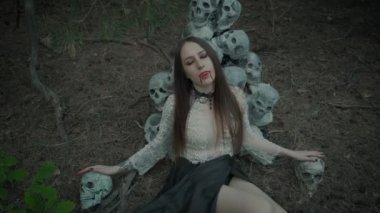 Genç bir vampirin portresi, yüzüne kan damlayan güzel kırmızı dudaklar, keskin sahte dişler. Karanlık bir gotik ormanda, insan kafatasının arka planına karşı bir model..