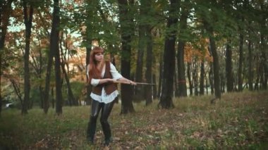 Ortaçağ kadın okçusu ağacın yanında saklanır ve yaydan ok atar. Adul fantezi kız klasik kostüm avcısı. Eski tarz bir savaşçı elinde silah tutuyor. Retro video görüntüleri. Sonbahar orman yaprakları.