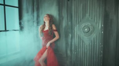 Zarif kırmızı gece elbisesi içinde genç ve güzel bir kadın kalenin eski odasının penceresi olan karanlık duvarın yanında duruyor. Manken pozu, gözler kapalı. Şık giyimli siluet. Seksi bacağı kesilmiş denizkızı.