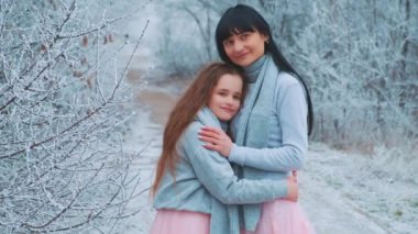 Mutlu aile annesi ve kızı fotoğraf çekimi için poz veriyor, küçük kızı sevgiyle kucaklıyor, iki insan birlikte. Kış ormanı ağaçları donar, beyaz kar yağar. Aynı giysiler gri kazak pembe elbise