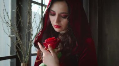 Güzel, genç, seksi, çekici bir kadın. Kırmızı kadife kapüşonlu, gotik karanlık odanın yanında duruyor. Arkaplan dalları. Kız gül yapraklarına hafifçe dokunuyor. Yüz makyajı, parlak bir akşam tatili.