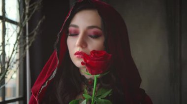 Gürültülü video. Kırmızı kadife kapüşonlu güzel ve çekici bir kadın. Nazikçe gül yüzüne dokun. Kızlar kokudan hoşlanır, aroma çiçeğinden. Pencere arkası gotik karanlık oda, ağaç dalları. Yakın plan portre.