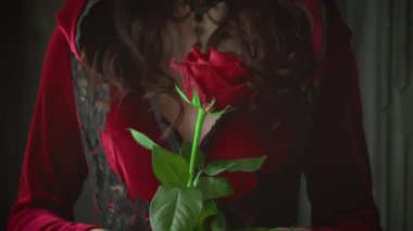 Gürültülü video. Yakın plan genç, güzel vampir kadın portresi. Ortaçağ kızgın cadısı elinde kırmızı bir gül tutuyor. Kadife seksi, klasik gotik elbise kapüşonu. Sanat cadılar bayramı makyajı. Gri gözler parlak dudaklar