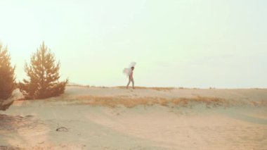 Yalnız efsanevi genç kutsal kutsal melek adam kumlu çölde yürüyor. Doğal afetler işte. Çevre koruma kavramı. Beyaz kanatlar, kuş tüyleri. Açık mavi gökyüzü, güneşli gün batımı, ağaçlar. Araba ayak izi.