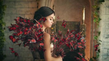 Güzel, görkemli, gizemli, genç kadın elf. Kız prenses ayakta duruyor, ortaçağ tuğla duvarlı antik şatosu. Birçok yapay kırmızı kelebekle süslenmiş yaratıcı, zarif bir sanat elbisesi.