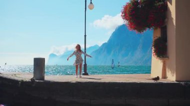 Neşeli, mutlu, genç bayan turist rıhtımda iyi eğlenceler. Dans eden kız ellerini kaldırdı. Kuaför elbisesi rüzgarı, doğanın tadını çıkar. Dalgalar mavi su, güneş gökyüzü. Lago di Garda Gölü. İtalya 'nın Riva Deltası.