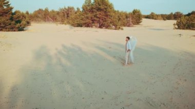 Hava atışı. Fantezi adam yalnız kutsal seksi melek kumlu çölde yürüyor. Yaratıcı kıyafet kostümü. Beyaz kanatlı kuş tüyü. Yaz doğası güneşli gün kumları, günbatımı ağaçları. Genç adam gökyüzüne bakıyor.