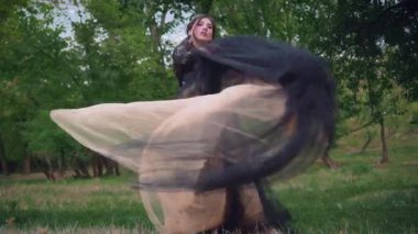 Karanlık bir ormanda siyah, yemyeşil, kaliteli uçan elbiseler içindeki gotik kraliçe. Dans eden kumaşlarda rüzgarda çırpınan kumaşlarda. Taçlı ve küpeli zarif göz alıcı saç stili. Arkaplan yeşili ağaçlar. Moda tasarımı