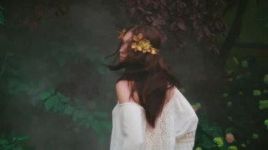 Esmer kız arkasını dönüp uzun saçlı sineğe bakıyor. Gülümseyerek mutlu bir kadın yüzü sallıyor. Arkaplan yeşil ağaç sisi bahar dumanı gibi. Yunan tanrıçası altın defne çelengi beyaz elbise. Fantezi Bayan Elf Kraliçesi