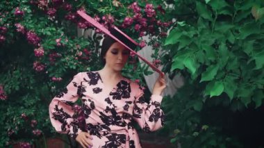 Eski film tarzı film tahılları eklendi. Harika bir kadın portresi. Kameraya poz veren geniş kenarlı şapka. Tanrıça modası. Pembe şık bir elbise. Yaz çiçekli ağaçlar çiçek açar. Çekici şirin yüz modeli