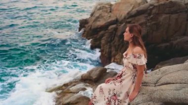 Gürültülü video. Genç kadın turist öfkeli mavi denizin taştan sahilinde oturuyor, dalgalar kayalara çarpıyor. Mutlu kız rüya görüyor, doğanın tadını çıkarıyor, Costa Brava sahilindeki parlak suyu görüyor Lloret de Mar.