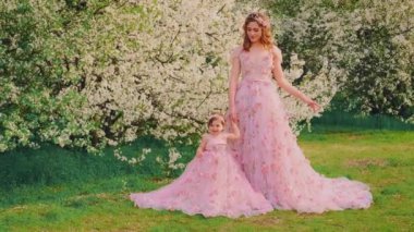 Aile fotoğraf çekimi için poz veriyor gülümseyen yüzlü kadın el ele tutuşuyor küçük kız. Mutlu anne ve kızı birlikte. Uzun pembe elbise. Kraliçe ve bebek prenses. bahar kiraz ağaçları beyaz çiçekler bahar doğası