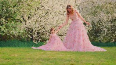 Aile fotoğraf çekimi için poz veriyor gülümseyen yüzlü kadın el ele tutuşuyor küçük kız. Mutlu anne ve kızı birlikte. Uzun pembe elbise. Kraliçe ve bebek prenses. bahar kiraz ağaçları beyaz çiçekler bahar doğası