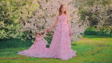 Aile fotoğraf çekimi için poz veriyor gülümseyen yüzlü kadın el ele tutuşuyor küçük kız. Mutlu anne ve kızı birlikte. Uzun pembe elbise. Kraliçe ve bebek prenses. bahar kiraz ağaçları beyaz çiçekler bahar doğası