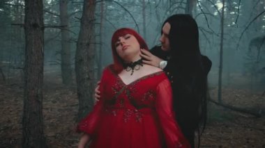 Siyah paltolu gotik vampir sarılıyor, ısırıyor, boynu öpüyor, soluk tenli, kırmızı dudaklı kadının kanını içiyor. Mutlu bayan fantezi çift iki kötü yüz. Kırmızı elbise. Mavi gizemli sis ormanı ağacı