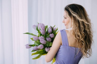 Lale buketi tutan mutlu sarışın kadın mor çiçekler yeşil beyaz pencere ışığı bırakıyor. Neşeli kız gülen surat dudaklar hediyeyi sever. Kadınlar Günü Tatili, 8 Mart, Bahar Konsepti Çiçek Kutlaması.