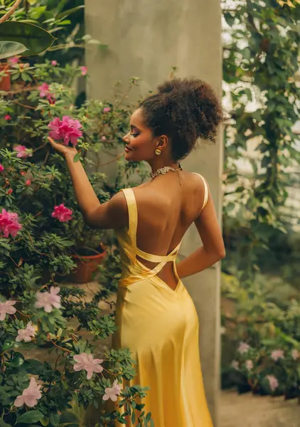 Sanat fotoğrafı gerçek insan fantezi kız eli taze pembe çiçeklere dokunuyor yeşil yaprak ağacı tropikal doğa. Mutlu Afrikalı Amerikalı güzel seksi kadın. Siyah kıvırcık saçlar sağlıklı cilt sarı elbise açık sırt