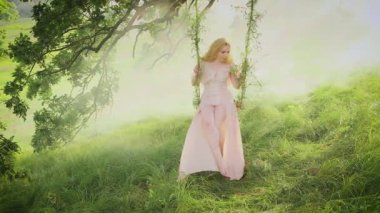 Fantezi kadın orman perisi salıncakta sallanıyor. Yaz doğası arka planı, beyaz sis dumanı, güneş ışınları, ilahi peri ışığı, yeşil ağaç, çimen. Klasik İpek Bej Elbise, Sarışın Uzun Saç