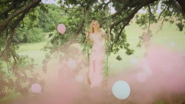 Genç mutlu sarışın kadın salıncakta sallanıyor. Saçlar ve uzun elbiseler rüzgarda uçuşuyor, hareket ediyor. Yaz doğası, yeşil çimenler, ağaç. Manken kız pozu. Dekorasyon pembe duman renkli hava balonları, top, kabarcıklar