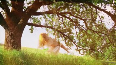 Genç, mutlu, neşeli, sarışın kadın salıncakta sallanıyor. Saçlar ve uzun elbiseler rüzgarda uçuşuyor, hareket ediyor. Yaz doğası, gün batımı, güneş ışınları, yeşil çimenler, ağaç. Kız model, gülen surat, olumlu duygular.