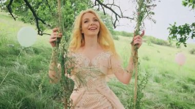 Doğal sarışın, uzun saçlı, mutlu genç bir kadının portresi. Şirin gülen surat, orman prensesi kız. Arka planda yaz yeşili bahar doğası. Manken pozu, elinde salıncak