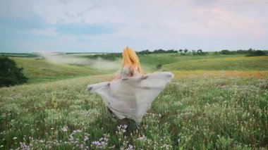 Genç mutlu sarışın kadın prenses yeşil bahar çiçekli tarlada yürüyor. Kız fırıl fırıl dans ediyor. Dalgalı etek, sallanan ipek mavisi gri elbise. Eski moda ortaçağ yaz kıyafetleri.