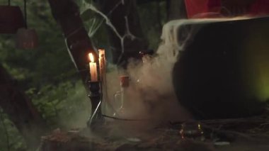 Simya ve iksirlerle ilgili sihirli bir video için ikinci çekim, ahşap masada cam şişelerin yanında yanan mum turuncu sıcak ateş cauldron, orman büyücüsü ve falcı beyaz sis içinde..