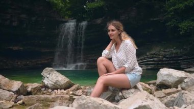 Çekici, sevimli sarışın kadın Georgia doğa dağlarının yanındaki tropikal göl şelalesinin yanında beyaz taşların üzerinde oturuyor. Günlük giysiler içinde mutlu bir turist sakin bir sessizlik ortamının tadını çıkarıyor. Gülümseyen şirin surat
